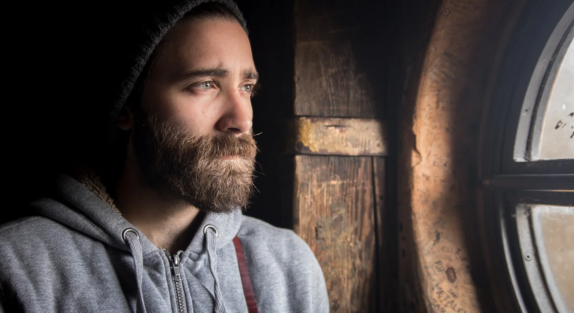 A man with a brown beard looking listfully out the window