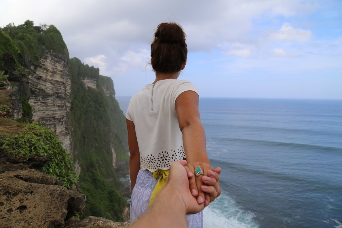 A lady holding hands with the camera person as she walks away from the camera towards a beach.