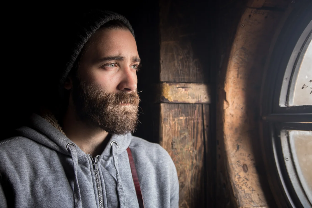 A man with a brown beard looking listfully out the window