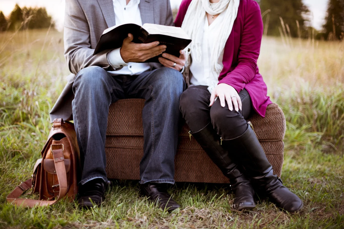 A man and a woman sitting on a log as the man reads to her from a book