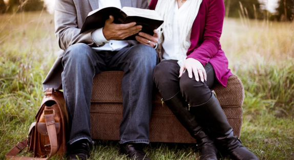 A man and a woman sitting on a log as the man reads to her from a book